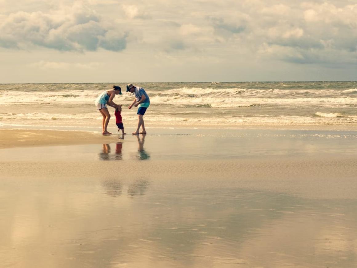 Two adults bending down to a small child at the edge of the sea on a wide, wet beach.